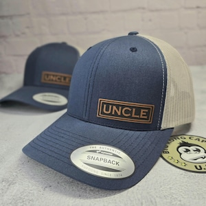 May include: A navy blue and white trucker hat with a brown leather patch that says "UNCLE". The hat has a mesh back and a YUPOONG SNAPBACK label.