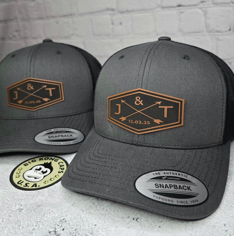 May include: Two gray and black baseball caps with a brown leather patch featuring the initials J & T and the date 12.02.25. The caps are both labeled "The Authentic Snapback Yupoong Since 1974".
