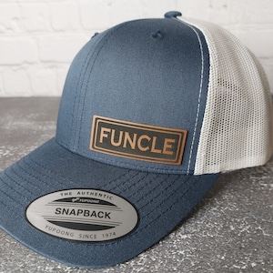 May include: A blue and white trucker hat with a brown leather patch that says "FUNCLE" on it. The hat has a mesh back and a label that says "The Authentic Yupoong Snapback Yupoong Since 1974".