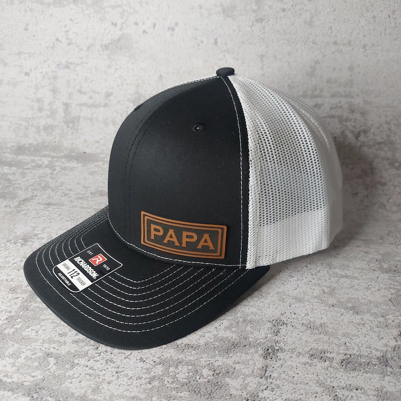 May include: Black and white trucker hat with the word "PAPA" on a brown leather patch. The hat has a curved brim with white stitching and a white mesh back. The Richardson brand tag is visible.