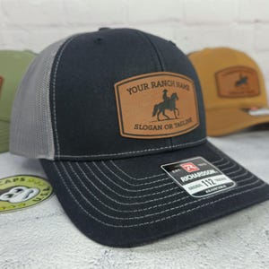 May include: Black and gray trucker hat with a leather patch featuring a rider on a horse and the text "YOUR RANCH NAME" and "SLOGAN OR TAGLINE". The hat has a curved brim and adjustable fit.