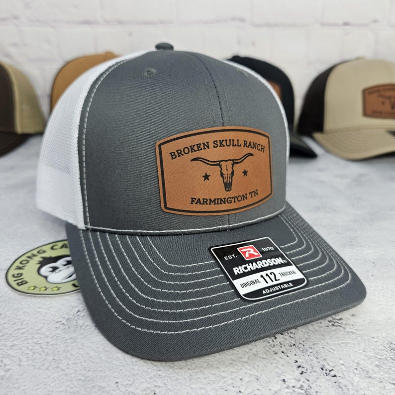 May include: Gray trucker hat with a white mesh back and a curved brim. The hat features a brown leather patch with the text "BROKEN SKULL RANCH FARMINGTON TN" and a bull skull graphic. The hat has a Richardson 112 tag.