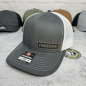 May include: Gray trucker hat with a white mesh back and a curved brim. The hat features the word "FREEDOM" on a rectangular patch. The Richardson brand label is visible on the brim. Other hats are visible in the background.