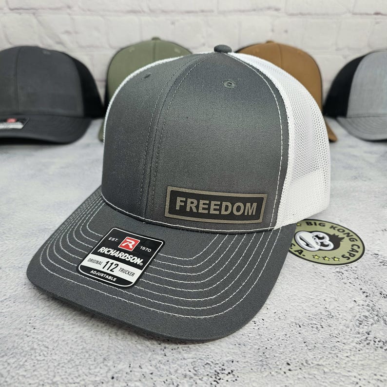 May include: Gray trucker hat with a white mesh back and a curved brim. The hat features the word "FREEDOM" on a rectangular patch. The Richardson brand label is visible on the brim. Other hats are visible in the background.
