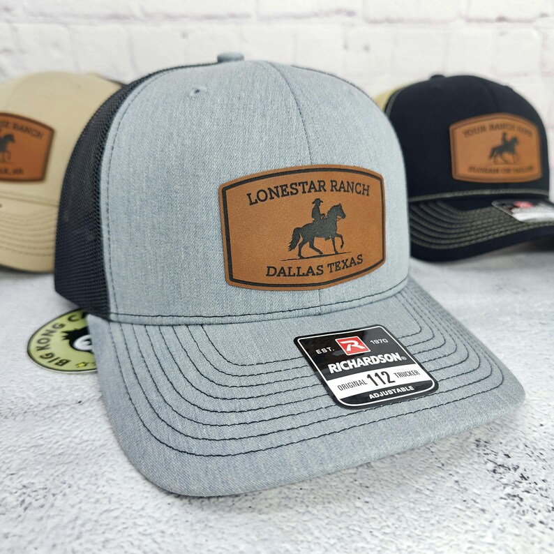 May include: A light gray trucker hat with a black mesh back and a brown leather patch. The patch reads "LONESTAR RANCH DALLAS TEXAS" with a silhouette of a cowboy on a horse. The hat is adjustable.