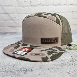 May include: A flat bill snapback hat with a camouflage pattern on the bill and a tan front panel. The hat features a brown leather patch with the word "DADDY" and a mesh back.