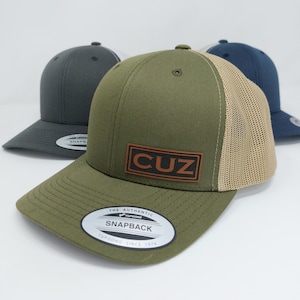 May include: A green and beige trucker hat with a brown leather patch that says "CUZ". The hat has a mesh back and a snap closure.