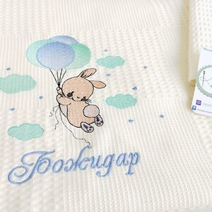 May include: A white waffle weave baby blanket with a light blue and green embroidered bunny holding balloons. The blanket has the text "Good night" embroidered in light blue.