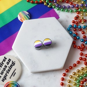 May include: A pair of non-binary pride flag earrings on a white hexagonal tile. The tile is surrounded by a rainbow beaded garland and a rainbow pride flag. There are also three buttons with pride-related messages.