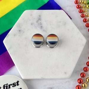 May include: A pair of rainbow flag-themed ear gauges. The gauges are made of stainless steel and have a silver finish. The rainbow flag design is printed on the front of the gauges.