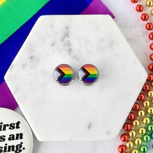 May include: Two silver ear gauges with a rainbow pride flag design featuring a black triangle in the center. The gauges are on a white marble surface.