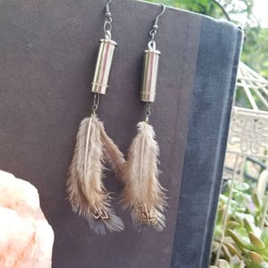 Può includere: Un paio di orecchini a forma di proiettile argentato con piume marroni che pendono dalla parte inferiore.