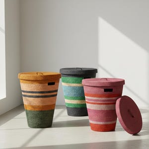 May include: Three cylindrical storage baskets with lids, each featuring horizontal stripes in distinct colour palettes: one in orange, yellow, and green; one in blue, green, and grey; and one in shades of pink and red, all with integrated handles.