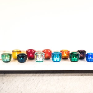 May include: A row of colourful glass candle holders in various shades of red, orange, yellow, green, blue, and purple. The candle holders are arranged in a line on a white surface.