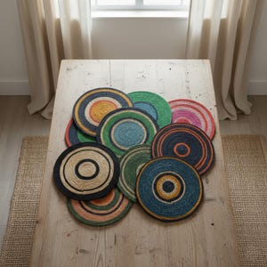 May include: A selection of circular jute placemats with concentric patterns in various colours, including black, blue, green, orange, and pink. The placemats are displayed on a light-coloured wooden table. Each placemat is approximately 30 cm in diameter.