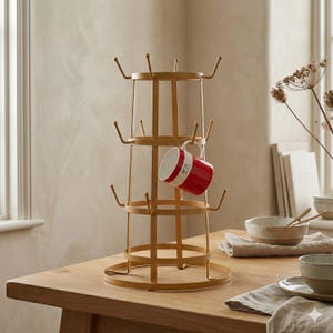 May include: A gold-coloured metal mug tree with a tiered, circular frame and multiple upward-curving hooks. A single red and white ceramic mug with the text N°29 printed on the side is hanging from one of the hooks on the middle tier of the stand.