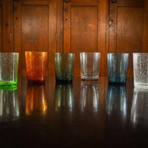 May include: Six clear glass tumblers with a textured surface. The tumblers are arranged in a row on a dark wooden surface. The tumblers are green, orange, brown, clear, blue, and white.