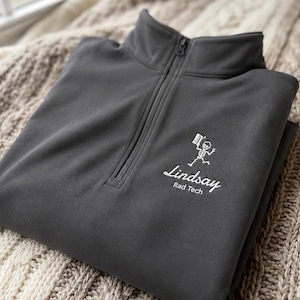 May include: A charcoal gray quarter-zip pullover with a white embroidered design featuring a skeleton holding a flag and the name "Lindsay" with "Rad Tech" underneath. The pullover is folded on a textured, neutral-toned surface.