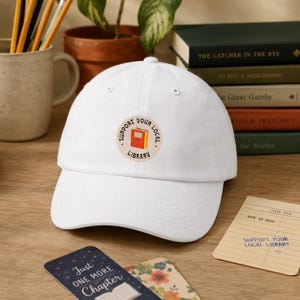 Peut inclure: Une casquette de baseball blanche avec un écusson circulaire sur le devant. L'écusson est beige clair avec une illustration de livre rouge au centre et le texte noir "SUPPORT YOUR LOCAL LIBRARY" courbé autour du livre. La casquette est structurée.