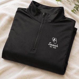 May include: Black quarter-zip athletic top with a mock neck collar. The shirt has a white logo with a heart, paw print, and stethoscope design, along with the name "Sarah" and the text "VET TECH".