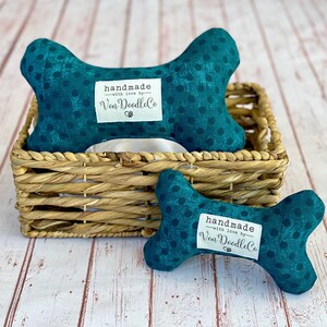 May include: Two teal blue dog bone toys with white polka dots. The larger bone is in a wicker basket. Both bones have a white label that reads "handmade with love by Van DoodleCo".
