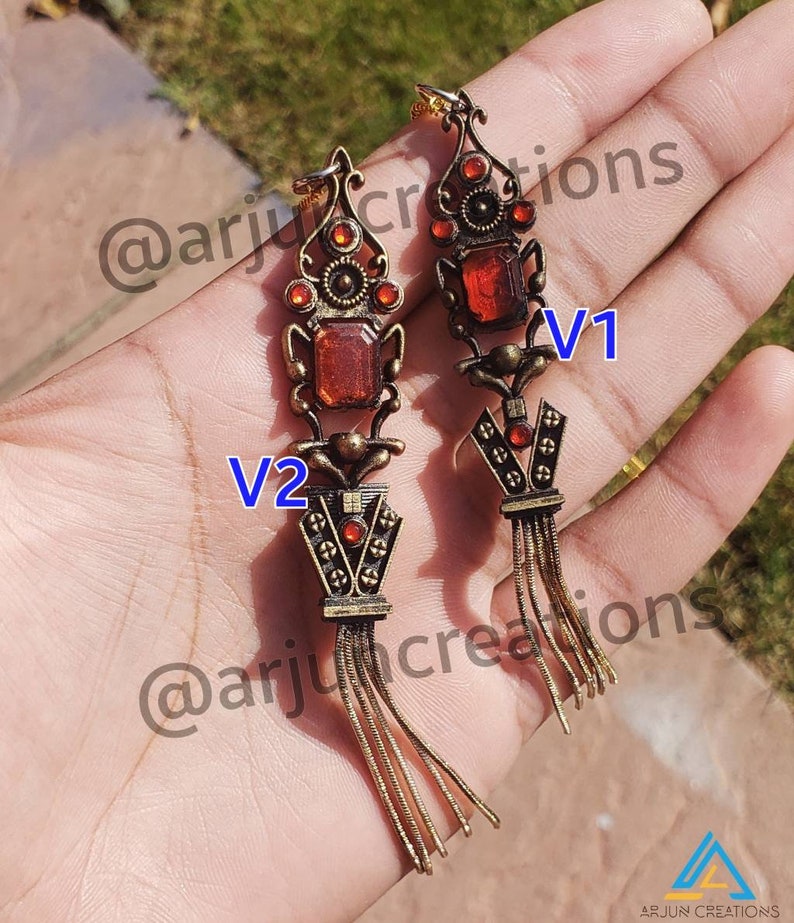 May include: A pair of gold-toned earrings with red gemstone accents and a tassel design. The earrings are labeled "V1" and "V2".