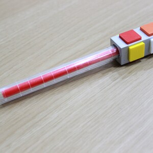 May include: A clear plastic tube with a red marker inside. The tube has a gray cap with a series of colorful square blocks on top. The blocks are red, orange, yellow, white, and yellow.