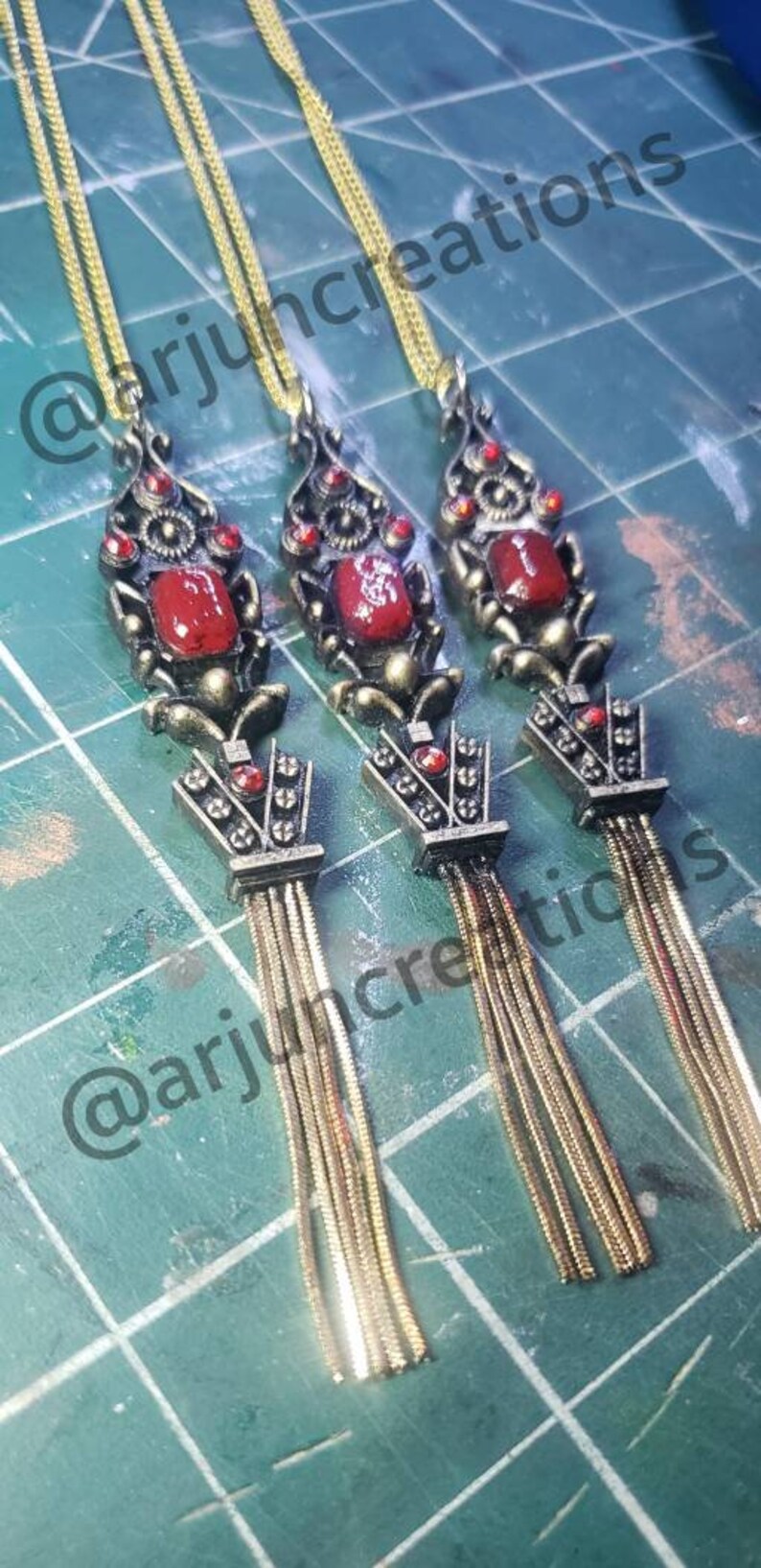 May include: Three gold necklaces with red gemstone pendants and tassel chains. The pendants are ornate and feature intricate designs. The chains are made of multiple strands of gold.