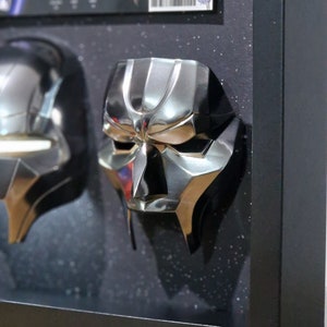 May include: Two silver-colored masks with a futuristic design are displayed in a black frame with a black background. The masks are mounted on a black surface. The frame is on a wooden surface.