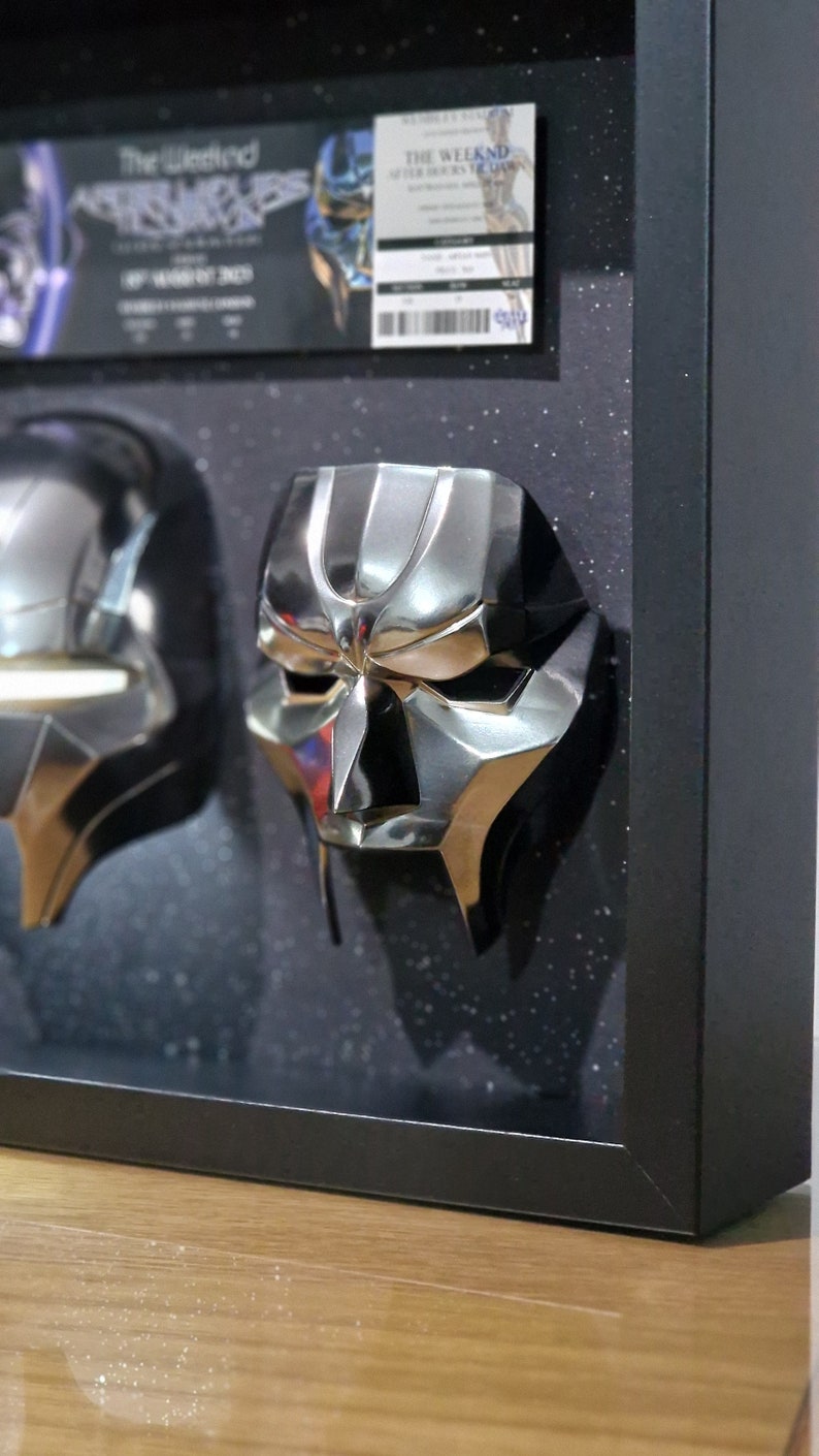 May include: Two silver-colored masks with a futuristic design are displayed in a black frame with a black background. The masks are mounted on a black surface. The frame is on a wooden surface.