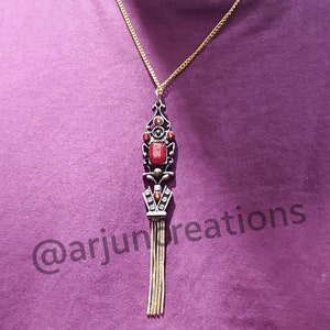 May include: A gold chain necklace with a red gemstone pendant. The pendant features a detailed design with a tassel. The necklace is on a purple shirt with the text "@arjuncreations".