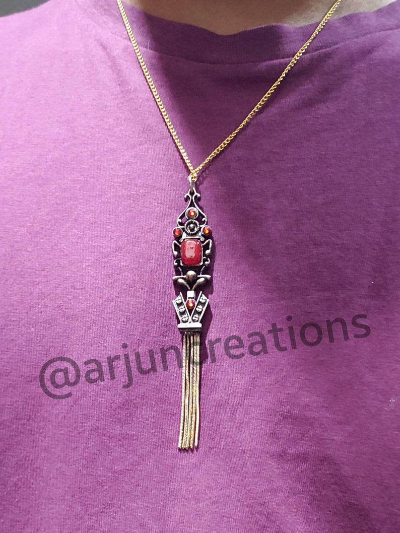 May include: A gold chain necklace with a red gemstone pendant. The pendant features a detailed design with a tassel. The necklace is on a purple shirt with the text "@arjuncreations".