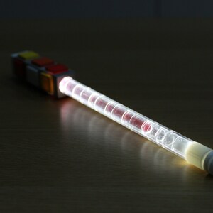 May include: A clear plastic tube with red and white accents, lit from within, creating a bright white glow. The tube is attached to a colorful block with red, yellow, and white squares.