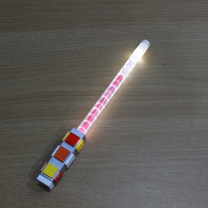 May include: A white light-up wand with a red, orange, yellow, and red block design at the base.