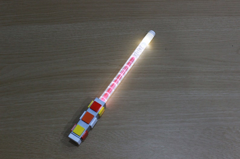 May include: A white light-up wand with a red, orange, yellow, and red block design at the base.