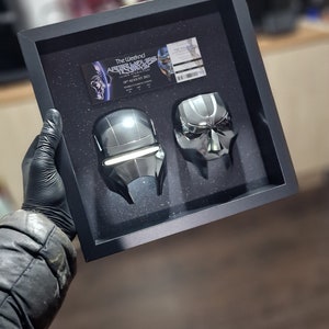 May include: A black framed display case with two silver metallic masks. The case has a ticket for "The Weeknd After Hours Til Dawn Tour" on August 18, 2022.