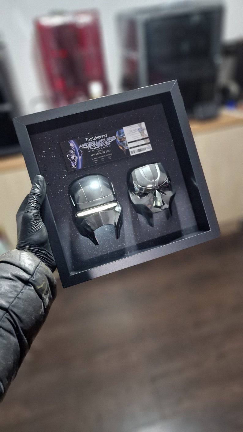 May include: A black framed display case with two silver metallic masks. The case has a ticket for "The Weeknd After Hours Til Dawn Tour" on August 18, 2022.