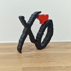 Op de afbeelding: Een zwarte 3D-geprinte sculptuur van "XO" met een rode hartaccent. De letters zijn verweven en hebben een getextureerd, geribbeld ontwerp. De sculptuur wordt getoond op een lichtbruin houten oppervlak.