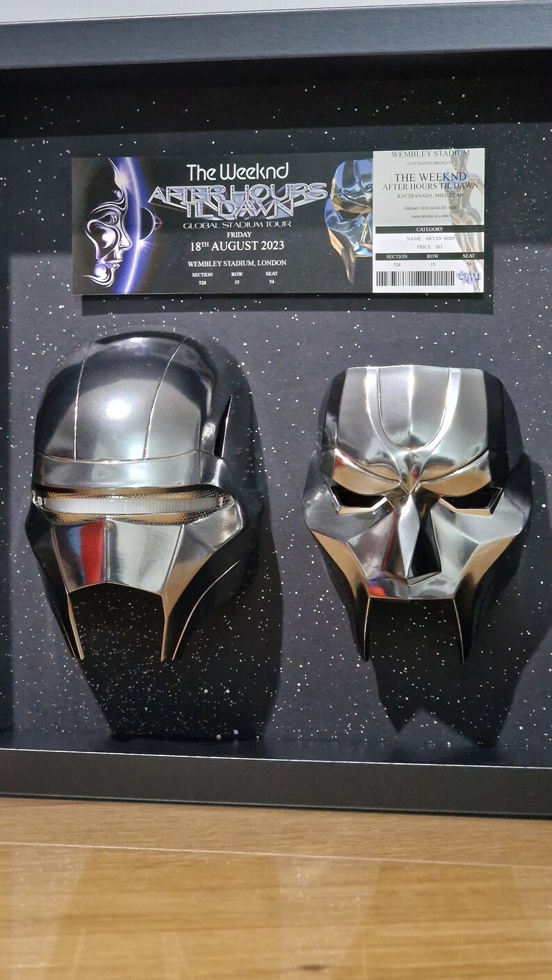 May include: Two silver masks, one with a visor and the other with a pointed chin, are displayed in a black frame with a sparkly background. The frame contains two tickets for The Weeknd's After Hours Til Dawn tour at Wembley Stadium on August 18, 2023.