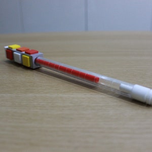 May include: A clear plastic pen with a red ink cartridge and a colorful Lego-like block attached to the top. The block is made of gray, yellow, red, and white plastic.