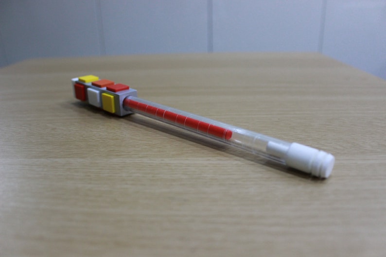 May include: A clear plastic pen with a red ink cartridge and a colorful Lego-like block attached to the top. The block is made of gray, yellow, red, and white plastic.