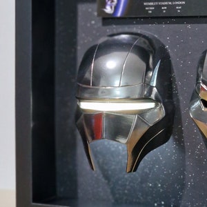 May include: A black framed display case with two silver helmets inside. The helmets have a futuristic design with a white light strip across the front. The case also includes a ticket for The Weeknd's After Hours Til Dawn tour in London on August 18, 2023.