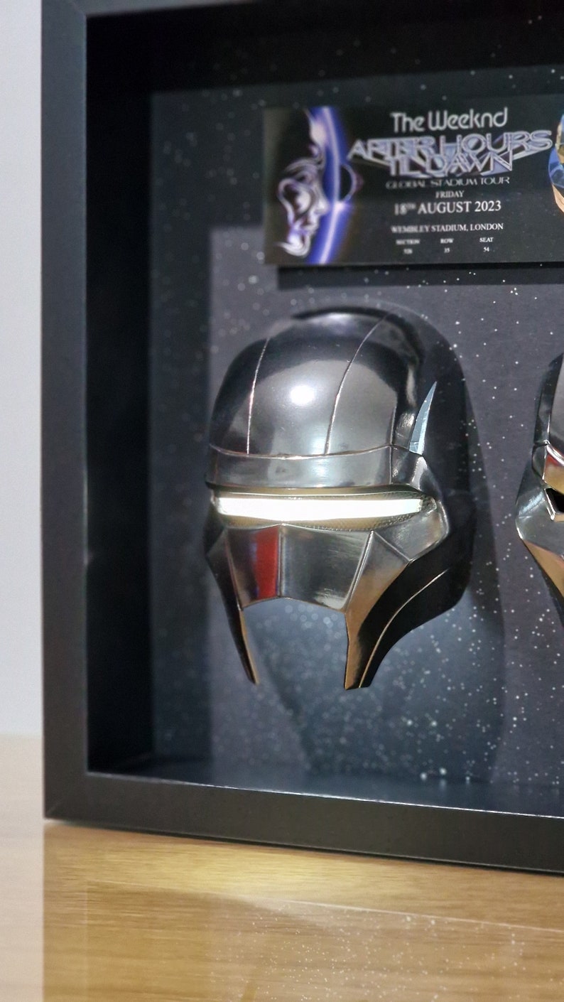 May include: A black framed display case with two silver helmets inside. The helmets have a futuristic design with a white light strip across the front. The case also includes a ticket for The Weeknd's After Hours Til Dawn tour in London on August 18, 2023.