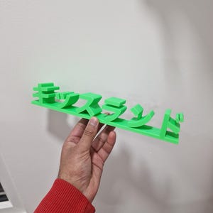 May include: A bright green, 3D-printed wall-mounted shelf featuring Japanese characters. The shelf is held by a person wearing a red long-sleeved shirt. The shelf is mounted on a white wall.