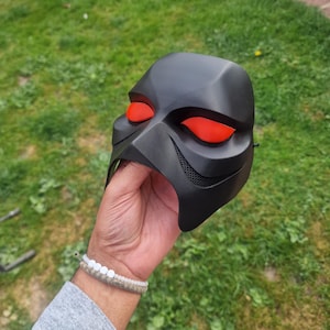 Op de afbeelding: Een zwart masker met rode oogkassen wordt omhoog gehouden tegen een groene grasachtergrond. Het masker heeft een strak, hoekig ontwerp met mesh-details bij de mond. De rode oogkassen vormen een opvallend contrast met het zwarte masker.