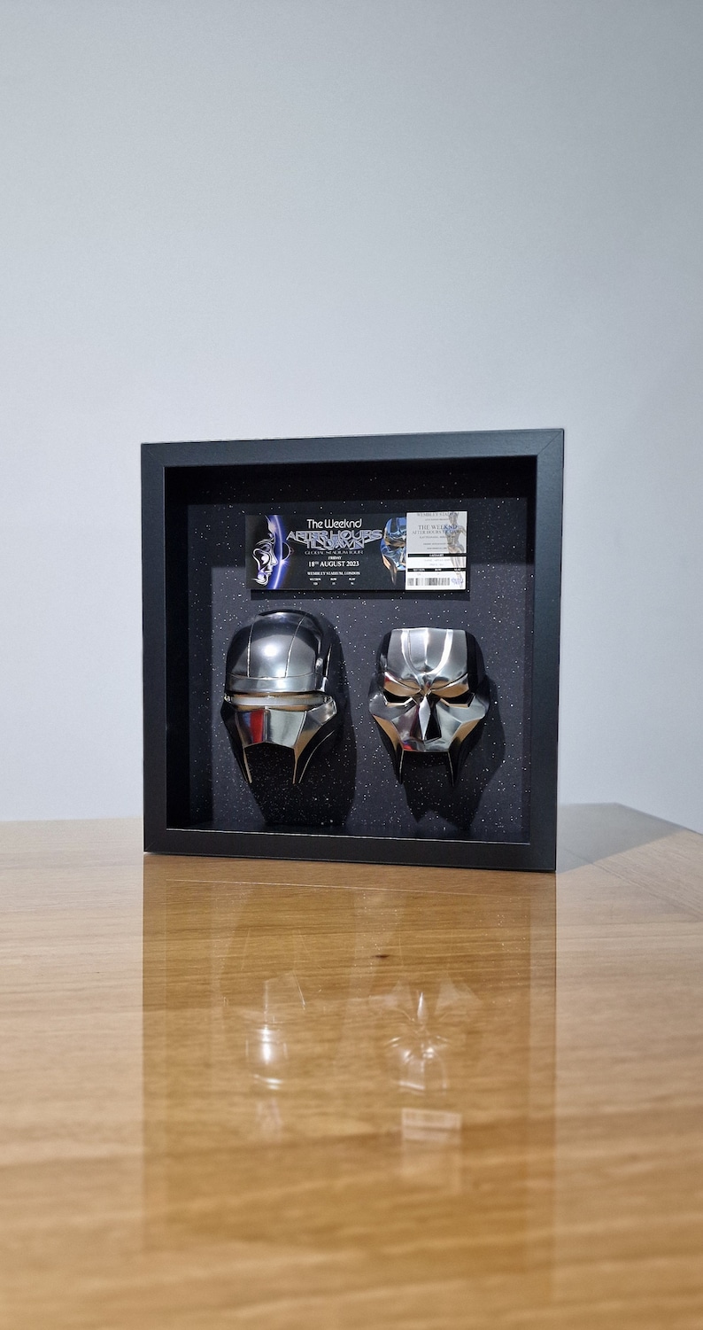 May include: A black framed display case with two silver helmets inside. The case has a ticket from The Weeknd's After Hours Tour on August 18, 2023, in London.