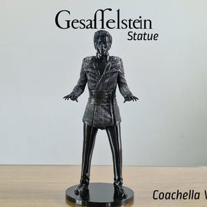 May include: A black statue of a figure in a textured jacket and trousers, standing on a round base. The statue is labelled "Gesaffelstein Statue Coachella Version" in black text.