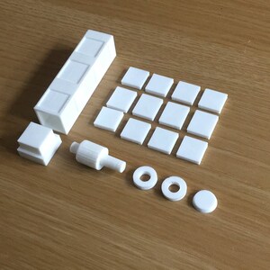 May include: A set of white plastic building blocks, including a long rectangular block, a small cube, a cylindrical piece, two rings, and a round disc. The blocks are arranged on a wooden surface.