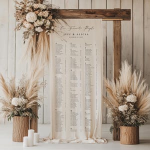 May include: A wedding seating plan featuring the text "Our Favorite People" and the names Jeff & Alicia. The cream-coloured fabric chart is displayed within a rustic wooden frame, adorned with floral arrangements and decorative pampas grass.