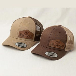May include: Two trucker caps with mesh backs and solid front panels. The tan cap features a brown leather patch reading A&M 8.15.26 BEST MAN. The brown cap features a brown leather patch reading A&M 8.15.26 GROOMSMAN. Both have a round sticker on the brim.
